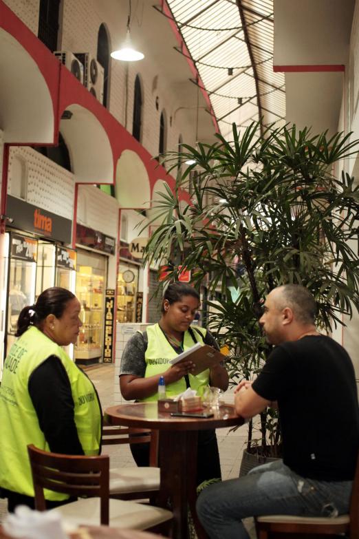 Agentes da Ouvidoria, identificadas por colete, conversam com um homem que está sentado a uma mesa de cafeteria. #pracegover