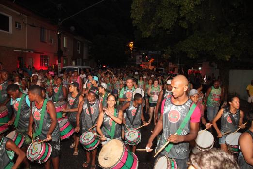 Integrantes de banda, com instrumentos de percussão e uniformizados estão na rua. #Pracegover