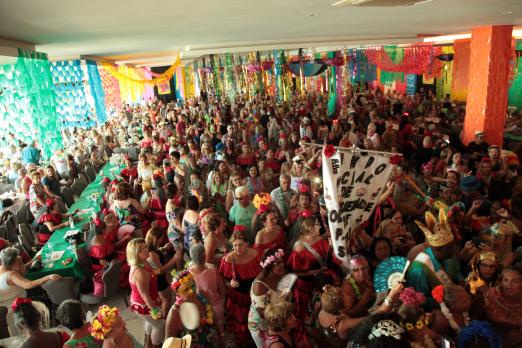 Salão de festas decorado para carnaval lotado de pessoas fantasiadas e dançando. #Pracegover