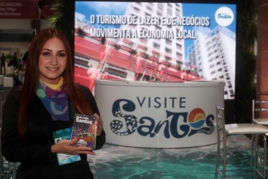 mulher segura panfleto em estande de santos de turismo #paratodosverem