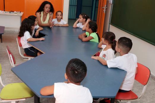 crianças pequenas, en torno de cinco anos, estão sentadas em torno de msa atentas à professora, que está na cabeceira, #paratodosverem 