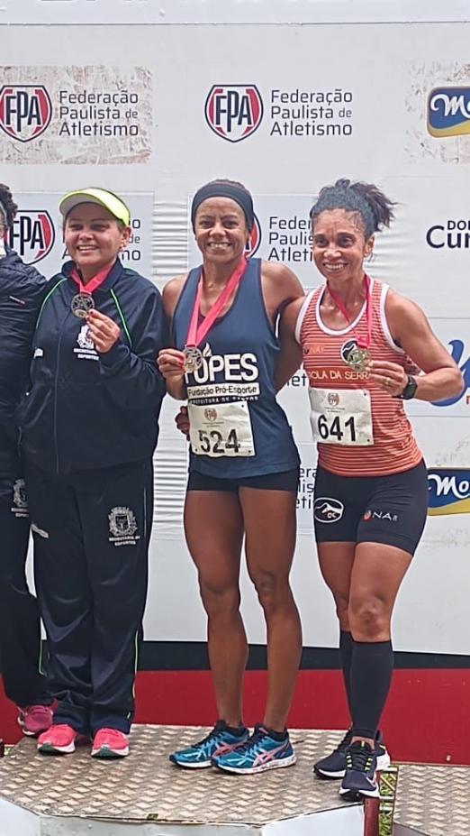 Três mulheres no pódio. A do centro é Ivanilda Pereira dos Santos, com a medalha no peito. #paratodosverem