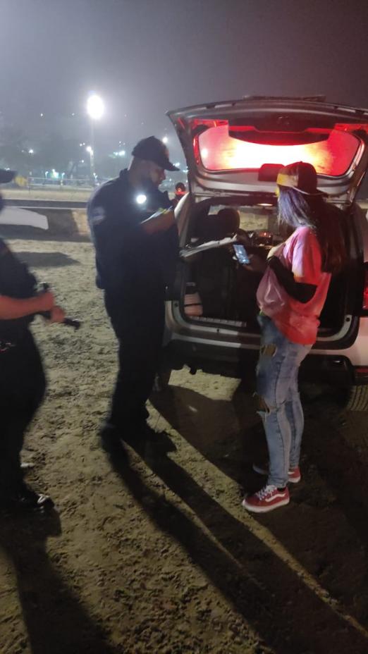 guarda municipal está na faixa de areia junto a uma mulher e um carro com o bagageiro aberto. Dentro há um equipamento de som. #paratodosverem