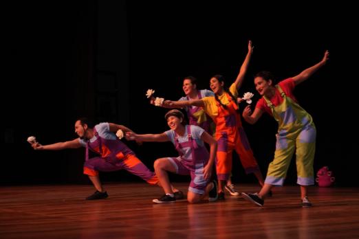 Artistas estão em palco de teatro, todos vestidos de palhaço e com flores nas mãos estendidas para a frente. #paratodosverem