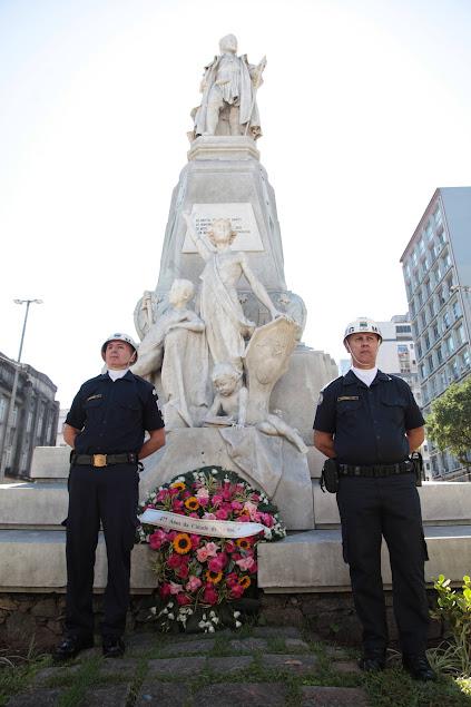 guardas municipais estão em pé diante de monumento a Braz Cubas e ao lado de coroa de flores. #paratodoverem