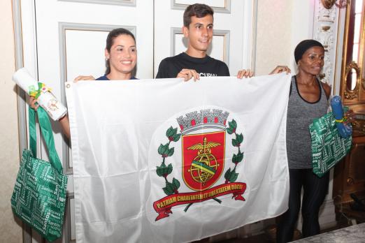 três corredores seguram bandeira com brasão de Santos #pracegover