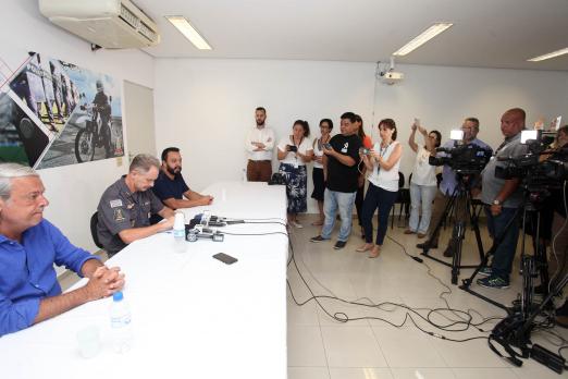 Três homens estão sentados a uma mesa. Sobre a mesa há microfones. Repórteres estão em pé diante dos homens que dão entrevista