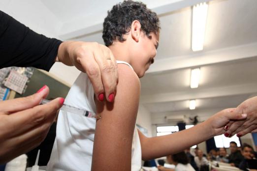 Mãos femininas aplicam injeção em braço de adolescente. O adolescente segura na mão de alguém. #Paratodosverem