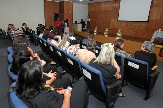 Pessoas sentadas em cadeiras de auditório prestam atenção em que está no palco, ao fundo. #Pracegover