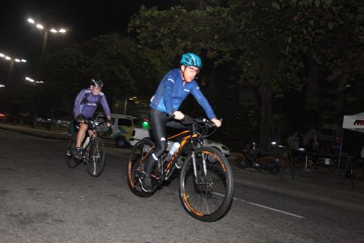 Ciclistas pedalam em avenida durante a madrugada. Na imagem aparecem dois. Eles usam uniforme e capacete. #Paratodosverem