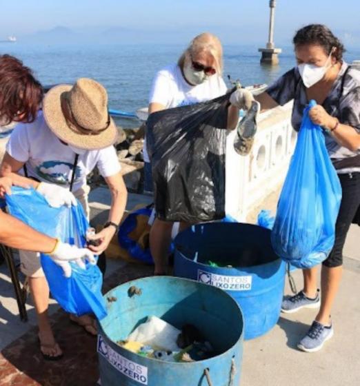 mulheres estão despejando lixo que está em sacos em toneis identificados pelo nome Santos Lixo Zero. #paratodosverem