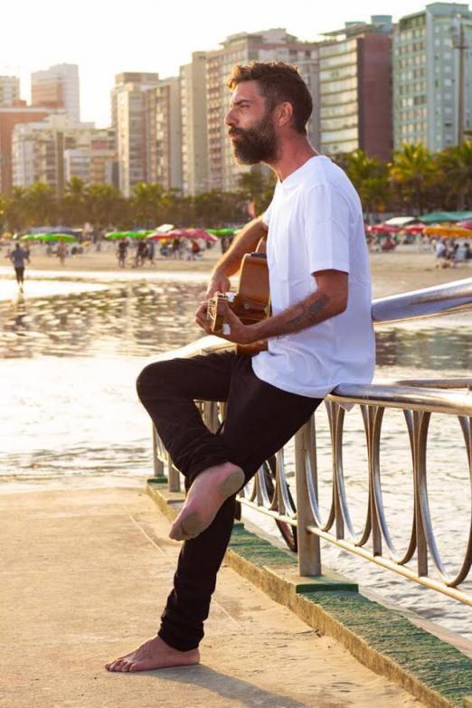 Lucas Real toca violão encostado no corrimão de ponte na praia. #Pracegover