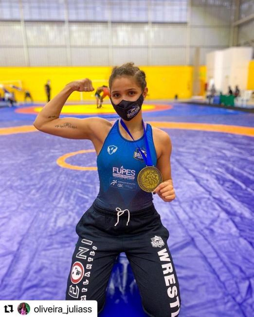 Julia está ajoelhada no tapete. Com a mão esquerda ela segura a medalha. O braço direito está flexionado para cima na altura do ombro, num gesto de força. #paratodosverem e a d
