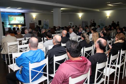 Público está sentado em cadeiras assistindo a uma apresentação ao fundo. #Pracegover