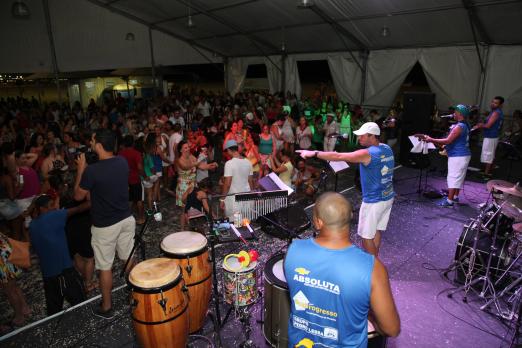 Baile de carnaval em tenda. No palco, os músicos se movimentam. Há instrumentos de percussão. #Paratodosverem