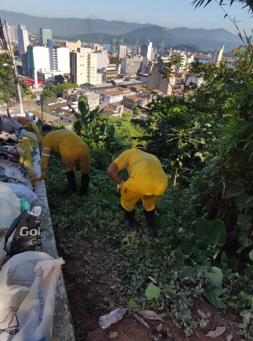 Homens trabalham em encosta do morro em área verde com lixo ao lado #paratodosverem