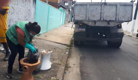 Agente de combate à dengue uniformizada e usando luva mexe em saco dentro de vaso sanitário deixado na rua. #paratodosverem