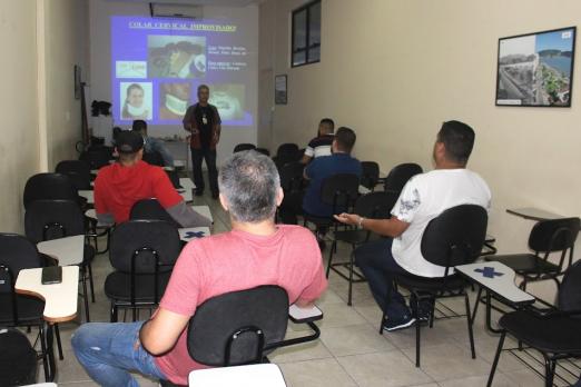 Pessoas estão sentadas em cateiras de uma classe. Um homem fala ao fundo diante de um telão. #paratodosverem