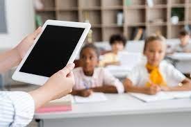 em primeiro plano, mãos seguram tablet. Ao fundo, crianças estão sentadas em sala de aula. #paratodosverem