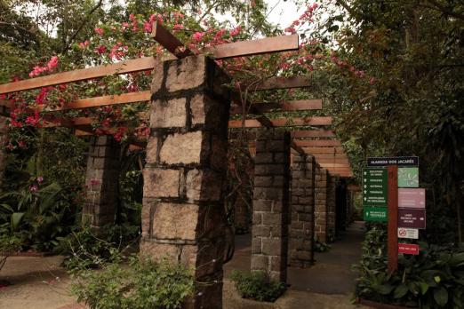 entrada do orquidário com pergolado coberto por vegetação e flores. Ao lado direito está afixada uma placa de orientação. #paratodosverem