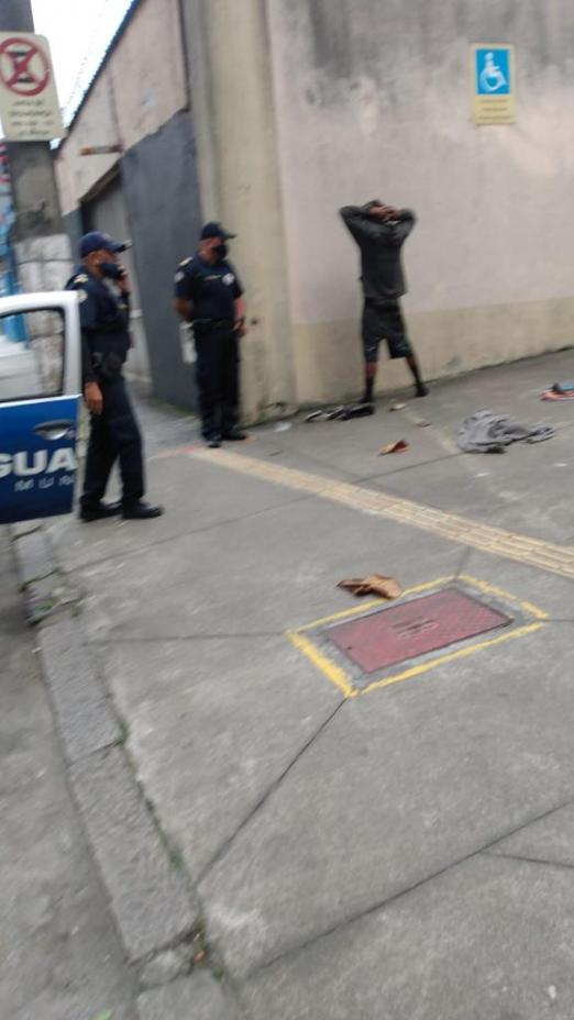 Dois guardas municipais estão ao lado de um homem de frente para a parede, com as mãos aos alto.#paratodosverem