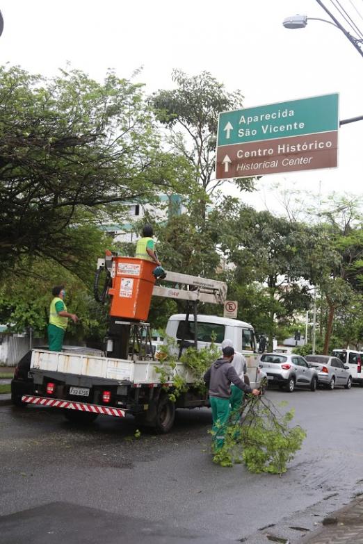 Caminhão está parado na rua. Um homem está no cesto basculante para fazer a poda de galhos de uma árvore. #Paratodosverem