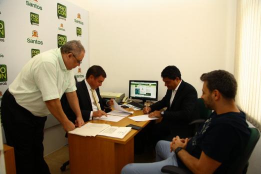 pessoas em torno de uma mesa. Banner na parede indica que estão no procon. #Paratodosverem