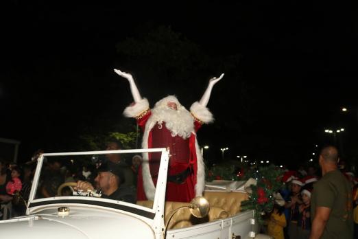 papai noel ergue os braços dentro de um carro aberto cumprimentado o público. O carro é dirigido por um motorista. É noite. #paratodosverem 