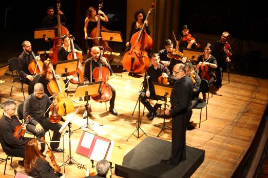 O grupo sinfônico de Santos em exibição em teatro. Os músicos estão dispostos no palco com seus instrumentos e diante de suas estantes com partituras. O maestro está à frente de todos sobre um púlpito. #Pracegover