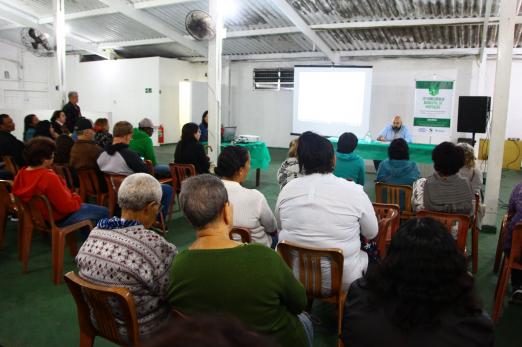 Salão com público sentado às cadeiras. As pessoas estão atentas a um homem que fala ao grupo. Ele está ao fundo diante de um painel. #Pracegover