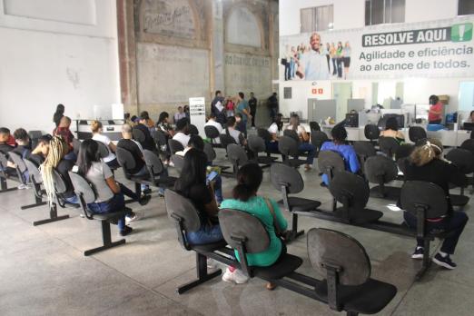 salão com várias cadeiras e pessoas sentadas. Ao fundo e afixada na parede há uma placa onde se lê Resolve Aqui. #paratodosverem