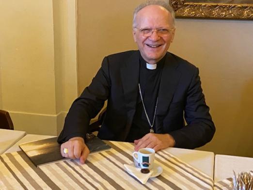 O arcebispo dom Angelo Vincenzo Zani está sentado a uma mesa. Há uma xícara de café na frente dele. #Pracegover