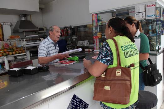 Agentes da Ouvidoria estão de costas para a foto e em frete a um balcão. Elas conversam com um homem atrás do balcão. #Pracegover