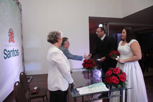 Casal de noivos, com as roupas tradicionais, está na frente de um casal atrás de uma mesa. Ao fundo um painel com o brasão de Santos. #Pracegover