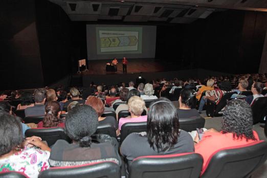 Teatro com cadeiras ocupadas majoritariamente por mulheres. Ao fundo, no palco, uma imagem está projetada em um telão. #Pracegover