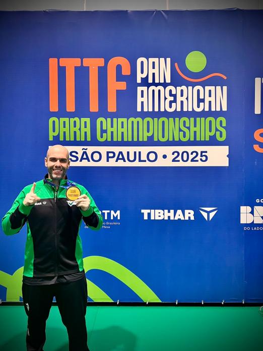 Mesatenista em pé junto ao painel do evento de tênis de mesa. #Paratodosverem
