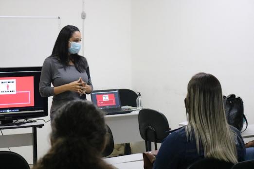 jovens estão sentados em sala de aula. Mulher está à frente de todos falando ao grupo. Há dois computadores atrás dela. - #paratodosverem