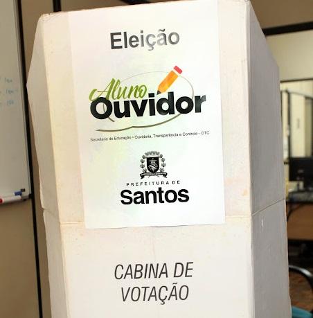Cabina de votação feita de papelão com cartaz do projeto. #pratodosverem