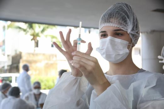 Mulher vestindo avental, usando touca e máscara manipula seringa de vacinação. #Paratodosverem