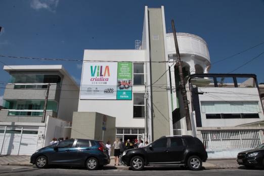 Fachada de vila criativa com placa no alto e carros estacionados na porta. #paratodosverem