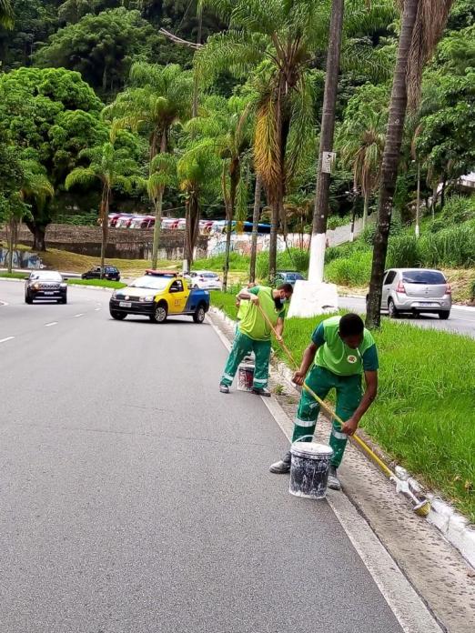 dois homens estão caiando guia junto a canteiro central. Eles seguram cabos e têm à frente latas com tinta. Ao fundo, um veículo da CET sinaliza a via. #paratodosverem