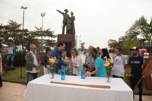 Celebração pela data no emissário submarino. Ao fundo está o monumento em homenagem à imigração japonesa no Brasil e, à frente uma mesa com vasos de flores e pessoas se cumprimentando. #Pracegover