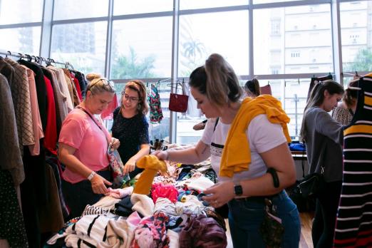 Bazar com mesa repleta de peças de roupas. Há mulheres examinando as peças. À esquerda há uma arara com várias roupas penduradas. #Pracegover