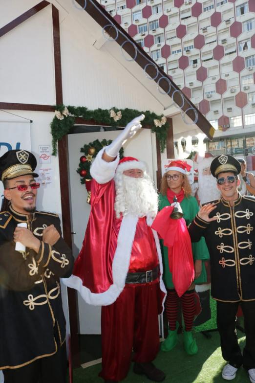 #pracegover Papai Noel acena ao píblico à frente de pequena casa montada na Praça Independência