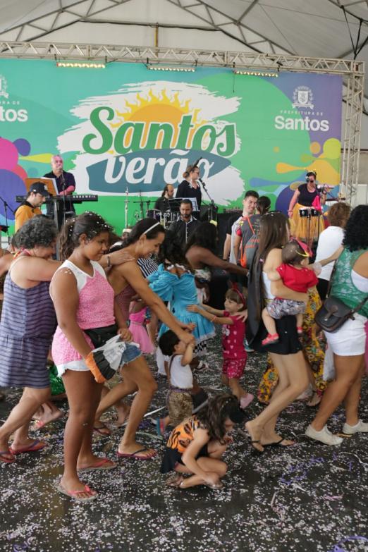 #pracegover Abaixo de tenda, crianças e adultos vrincan enfileirados á frente de palco e sobre chão repleto de confetes