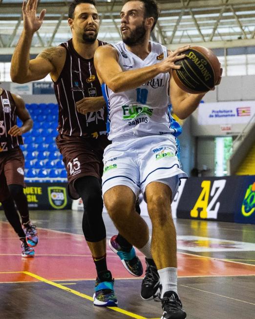 atleta segura bola no jogo enquanto outro faz marcação #paratodosverem