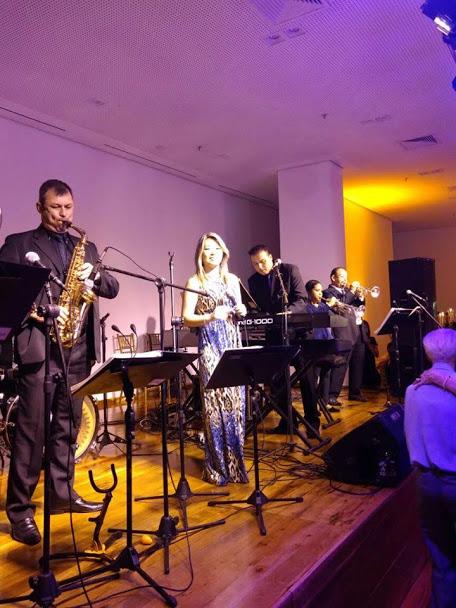 Músico toca saxofone, no primeiro plano, ao lado de cantora e outros integrantes da banda. #pracegover