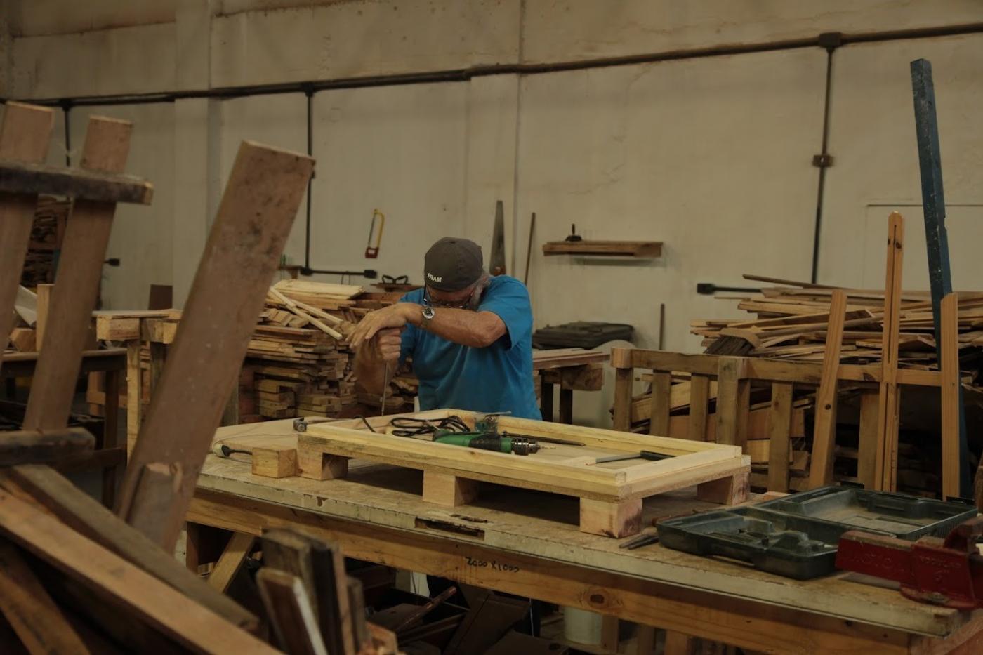 homem está fazendo trabalhos de marcenaria em meio a vários pedaços de madeira. #paratodosverem 