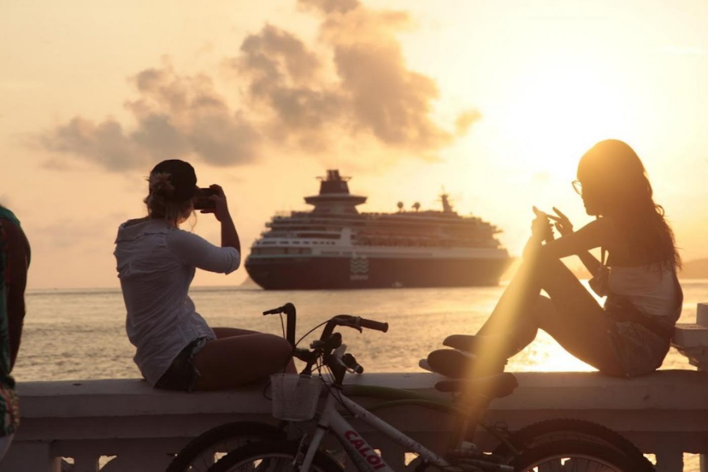 duas pessoas estão sentadas na mureta da orla assistindo à saída de um navio. Os dois fotografam com celulares. A foto foi feita ao por do sol, que cai e brilha do lado direito. #paratodosverem