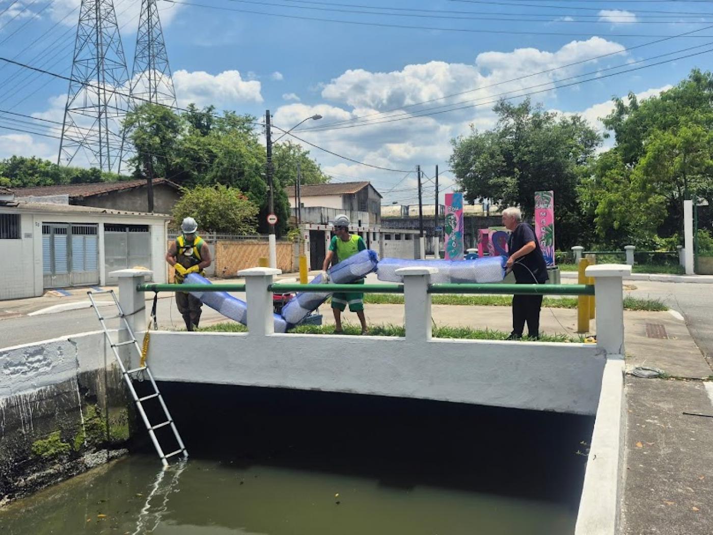 homens estão do lado de fora do canal segurando barreira e preparando para colocar na água. #paratodosverem 